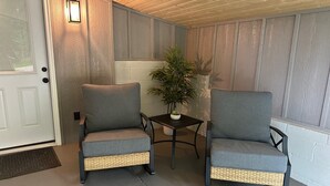 To grå lænestole med flettede sæder, et lille bord og en potteplante på en overdækket veranda med trævægge og en hvid dør.
