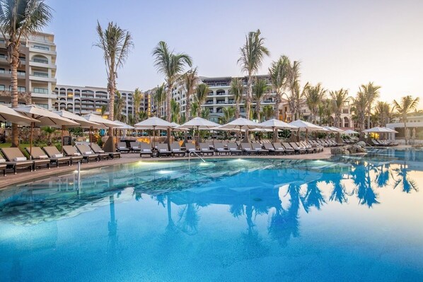 A pool area with lounge chairs and umbrellas, surrounded by palm trees and hotel buildings.