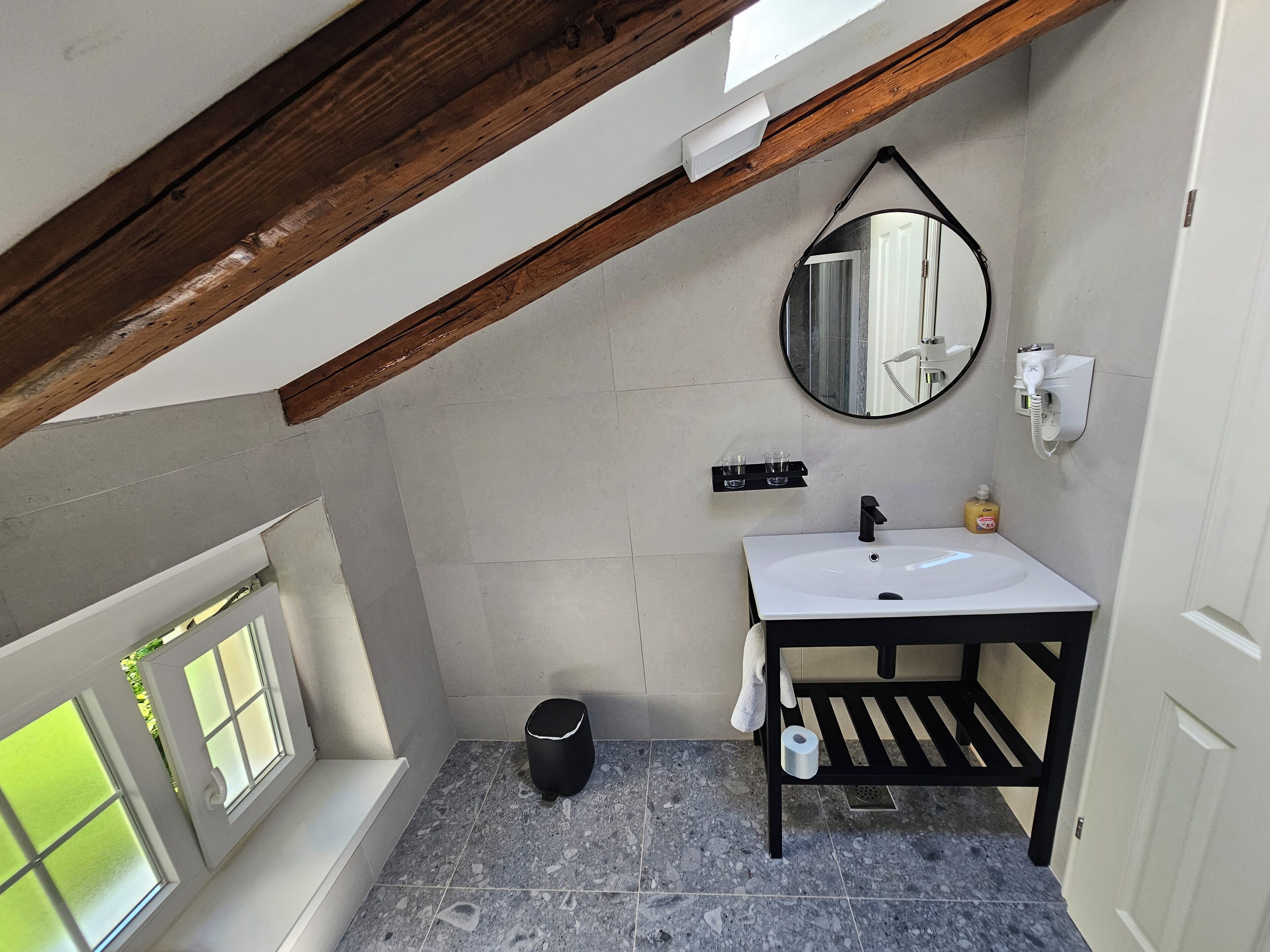 A small, modern bathroom with a white sink, a mirror, and a black vanity. There is a window with frosted glass and a view of greenery outside.