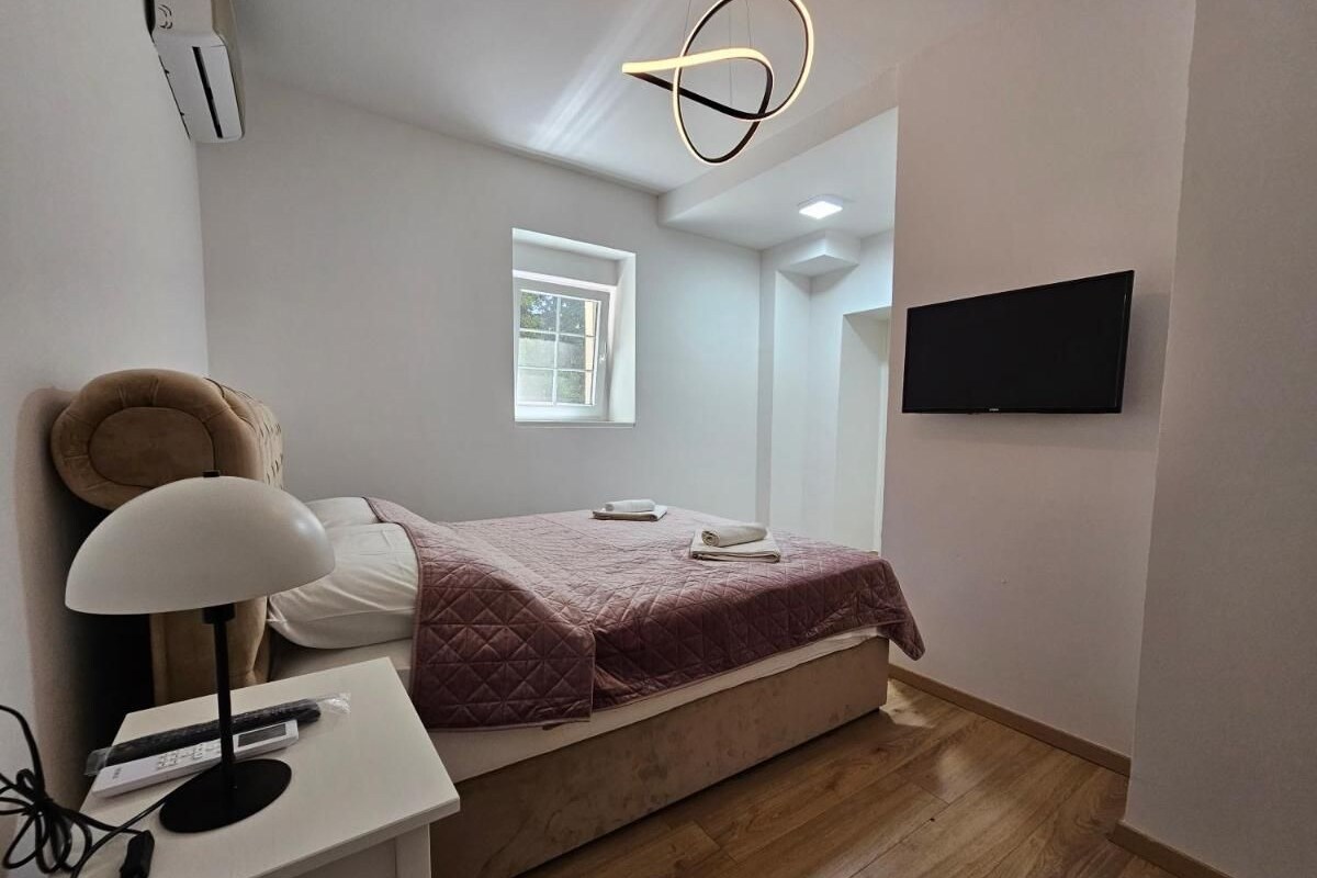 A bedroom with a bed, a bedside table with a lamp, a TV mounted on the wall, and a window.