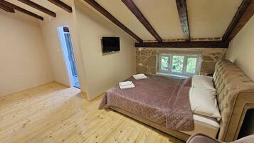 A bedroom with a wooden floor, a bed with a quilted cover, a stone wall, and a window with a view of greenery.