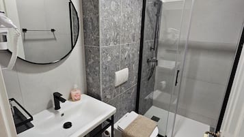 Un baño moderno con un lavabo blanco, un espejo, un secador de pelo de mano, una cabina de ducha de vidrio y una toalla en el inodoro.