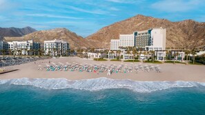 A beachfront hotel with multiple buildings, a sandy beach with umbrellas, and mountains in the background. - Seafront 4BR Apartment with Assistant Room in The Address Fujairah Residences Block B, Fujairah (Fujairah)