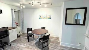 Un comedor con una mesa redonda y cuatro sillas, una pequeña cocina con mostrador y armarios, y un espejo grande en la pared.