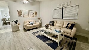Ein Wohnzimmer mit beiger Couch, Couchtisch und Deckenventilator. An den Wänden hängen Bilderrahmen und es gibt ein Fenster mit Jalousien.