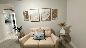 Ein Wohnzimmer mit einem beigefarbenen Sofa, einem kleinen runden Tisch mit einer Pflanze und drei gerahmten Bildern an der Wand.