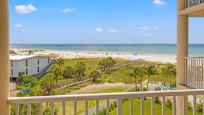 A beachfront view from a balcony, including a sandy beach, ocean, and a building with a staircase leading down to the beach.