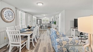 A modern kitchen with white cabinetry, a dining area with a wooden table and white chairs, and a patterned sofa. - 5-Bedroom Large Home in Windy Hill -Jus' Yungins (North Myrtle Beach)