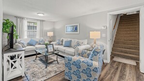 A living room with a white sofa, a patterned armchair, a glass coffee table, and a TV on a white stand. - 5-Bedroom Large Home in Windy Hill -Jus' Yungins (North Myrtle Beach)