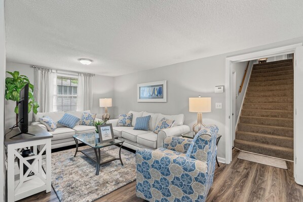 A living room with a white sofa, a patterned armchair, a glass coffee table, and a TV on a white stand. - 5-Bedroom Large Home in Windy Hill -Jus' Yungins (North Myrtle Beach)