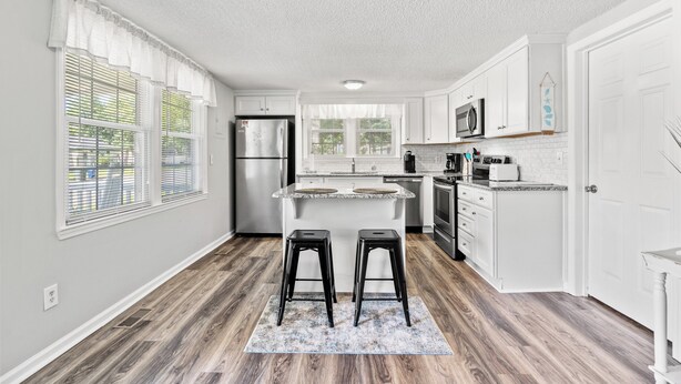 A modern kitchen with white cabinets, a central island, stainless steel appliances, and a window with blinds. - 5-Bedroom Large Home in Windy Hill -Jus' Yungins (North Myrtle Beach)