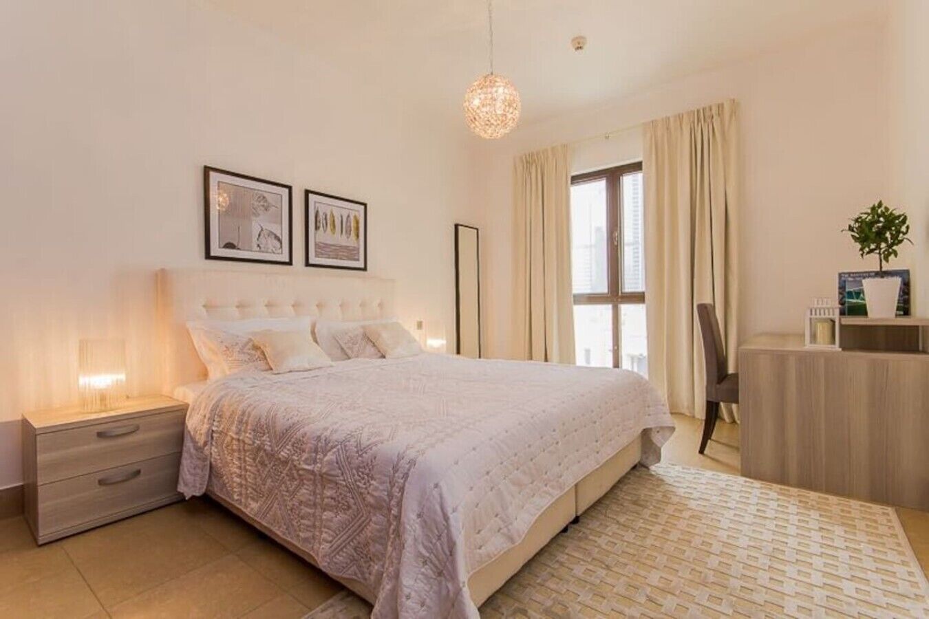 A bedroom with a bed, bedside tables, a desk, a chair, a hanging light fixture, and a window with curtains.