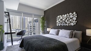 A modern bedroom with a large bed, a desk, and a chair, a potted plant, and a city view.