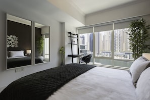 A modern bedroom with a large bed, a city view, a black chair, and a mirror reflecting the bed.
