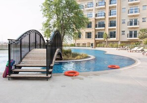 A swimming pool area with a wooden deck, a metal bridge, and a building in the background.