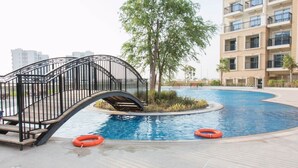 A swimming pool with a bridge, surrounded by a building with balconies and a tree.