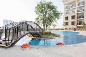 A swimming pool with a bridge, surrounded by a building with balconies and a tree.