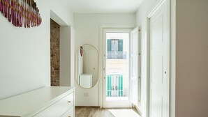 Un couloir avec une commode blanche, un miroir rond et une vue sur un balcon par une porte ouverte.