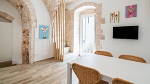 Un intérieur moderne avec des murs en pierre apparentes, une table blanche, des chaises en osier et un téléviseur à écran plat fixé au mur.