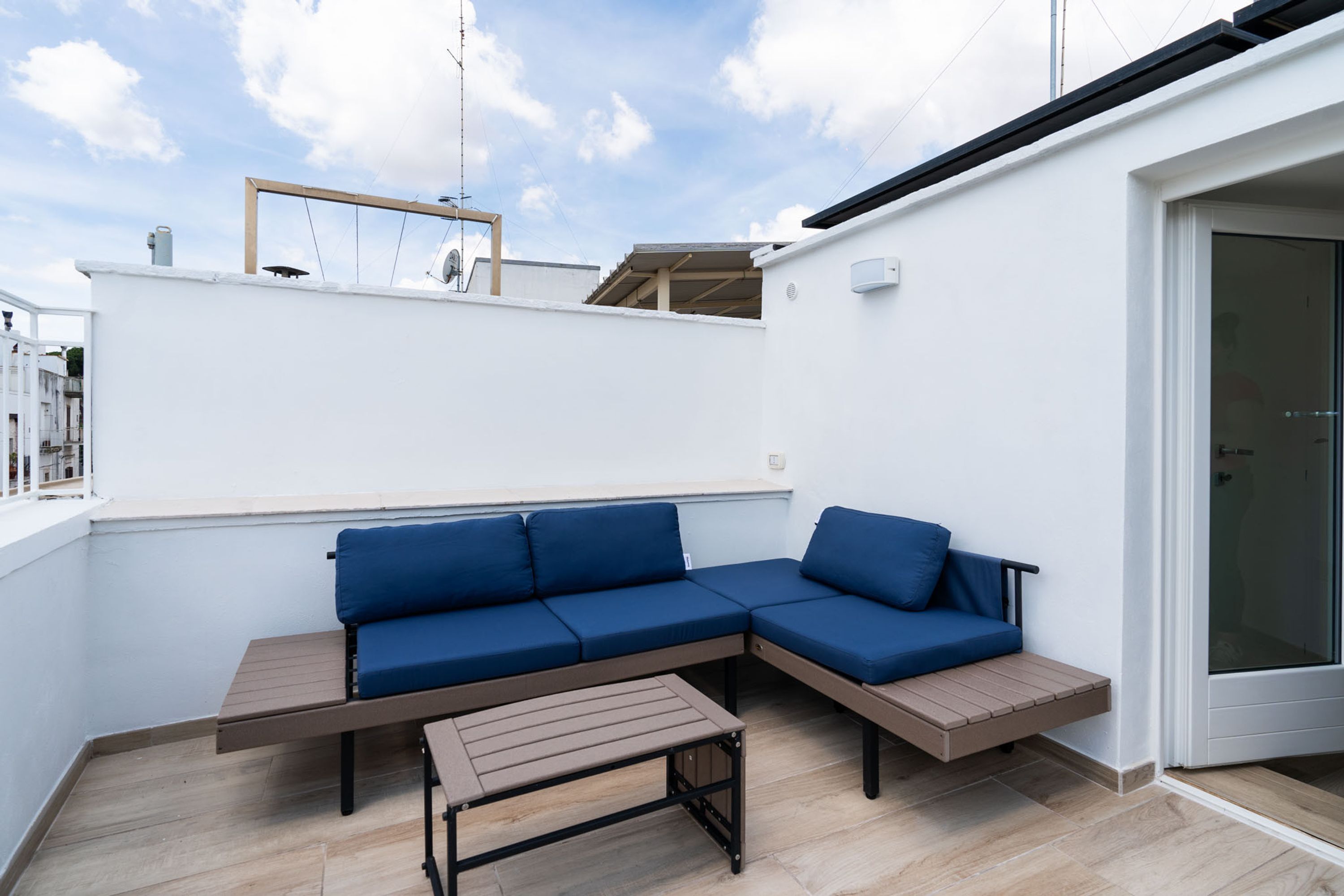 Un espace extérieur moderne, aménagé avec un canapé bleu et une table en bois, situé à côté d’un mur blanc et d’une porte vitrée.
