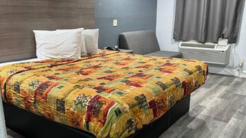 Quarto de hotel com uma cama grande, uma poltrona cinzenta, uma mesa de cabeceira e um aparelho de ar condicionado fixado na parede.