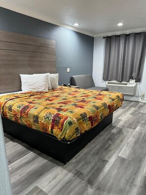 A hotel room with a large bed, a grey armchair, a bedside table, and a wall-mounted air conditioning unit. - LUXURY INN (Houston)