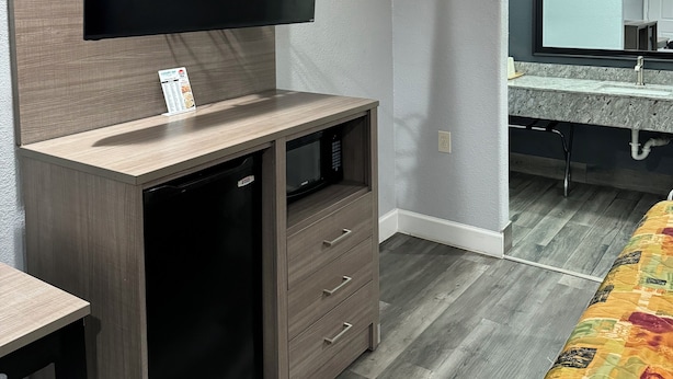 A compact living space with a flat-screen TV mounted on a wooden cabinet, a built-in microwave, and a small refrigerator. A bathroom with a vanity and mirror is visible through an archway. - LUXURY INN (Houston)