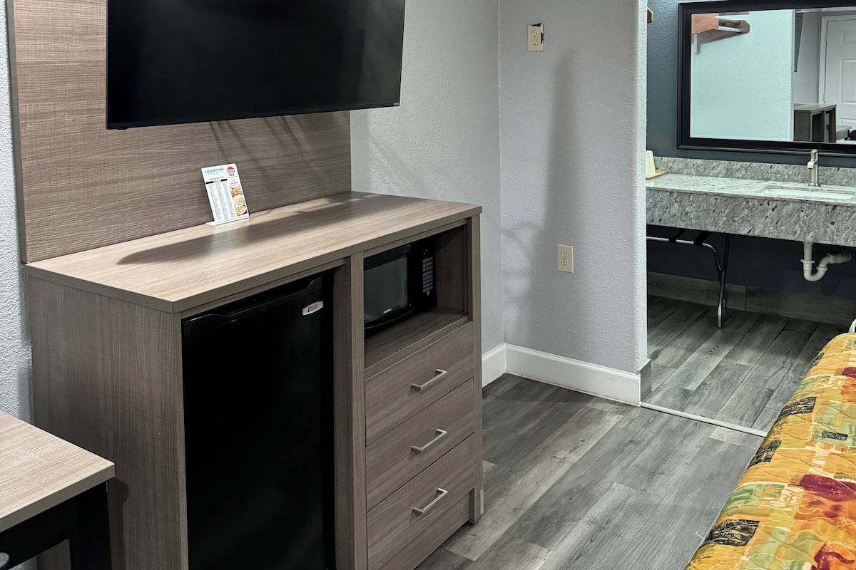 A compact living space with a flat-screen TV mounted on a wooden cabinet, a built-in microwave, and a small refrigerator. A bathroom with a vanity and mirror is visible through an archway.