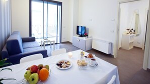 Ett modernt vardagsrum med en soffa, ett matbord med frukt och bakverk, en tv och ett stort fönster.