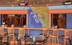 Un restaurant orné d’une grande fresque représentant la côte du golfe, avec des chaises et des tables en bois, et un bar où sont exposées des bouteilles et des verres.