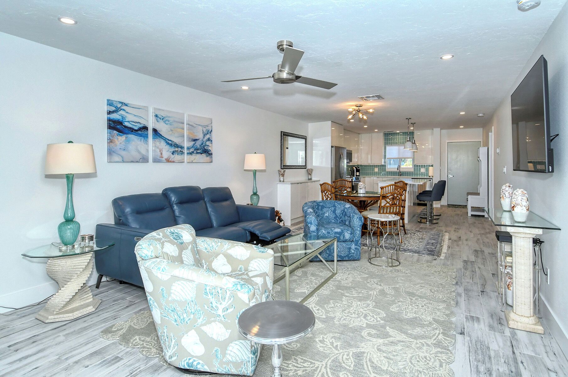 A modern living room with a blue sofa, patterned armchair, and glass coffee table. A ceiling fan and recessed lighting are visible.