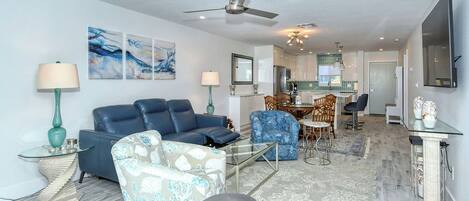 A modern living room with a blue sofa, patterned armchair, and glass coffee table. A ceiling fan and recessed lighting are visible.