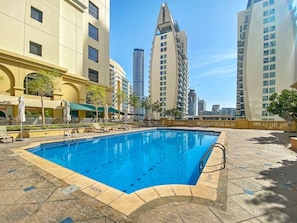 Une piscine dotée d’une terrasse carrelée, entourée d’immeubles avec des balcons, sous un ciel dégagé.
