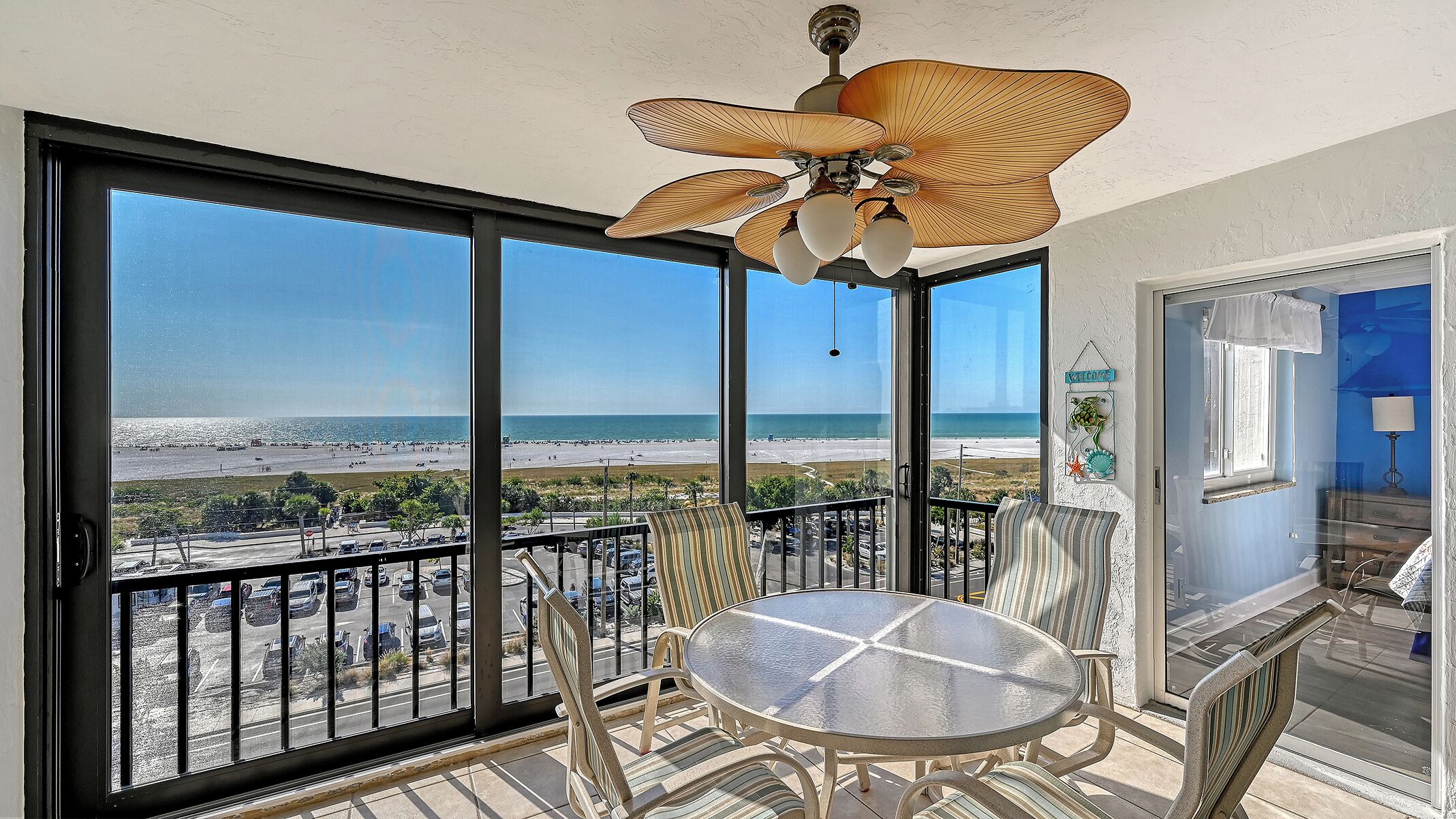 A balcony with a glass table and chairs, a ceiling fan, and a view of the beach.