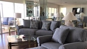 Ein modernes Wohnzimmer mit Ecksofa und Hocker, einem Glastisch und einem Fernseher im Hintergrund.