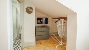 Une petite pièce avec un plancher en bois, une commode grise, une horloge murale, un téléviseur et un étendoir à linge blanc.