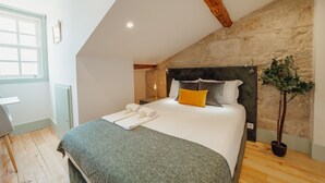 Une chambre avec un mur en pierre, un lit avec une literie blanche et grise, un plancher en bois et une plante en pot.