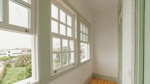 Ein Zimmer mit drei großen Fenstern, Holzdielenböden und Blick auf Gebäude und Grünflächen.