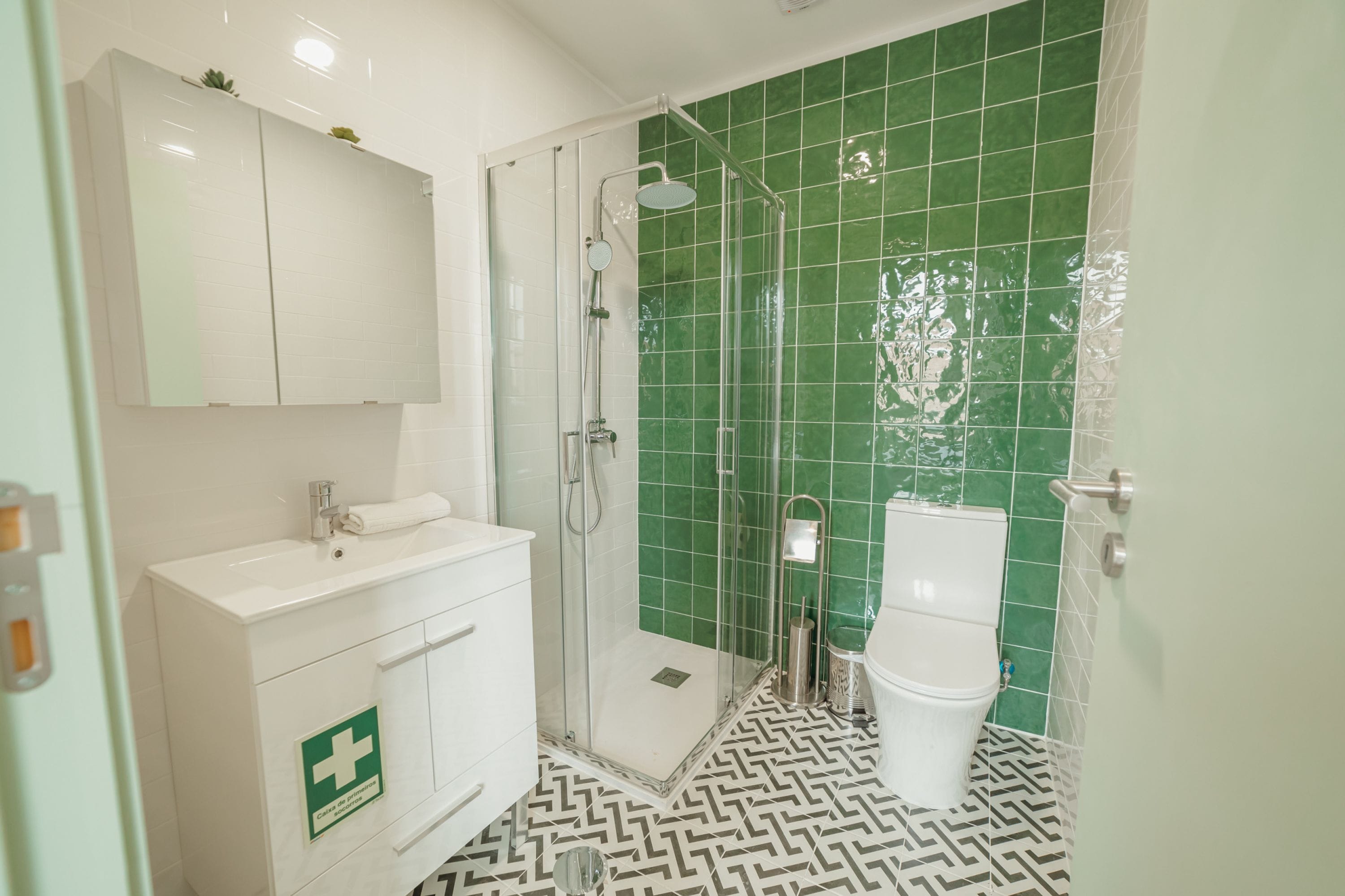 Une salle de bain avec un lavabo blanc, une douche en verre, des toilettes et des murs recouverts de carreaux verts.