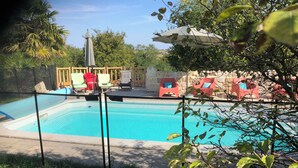 Ein Schwimmbecken mit Holzterrasse, umgeben von Grünpflanzen und roten Stühlen.