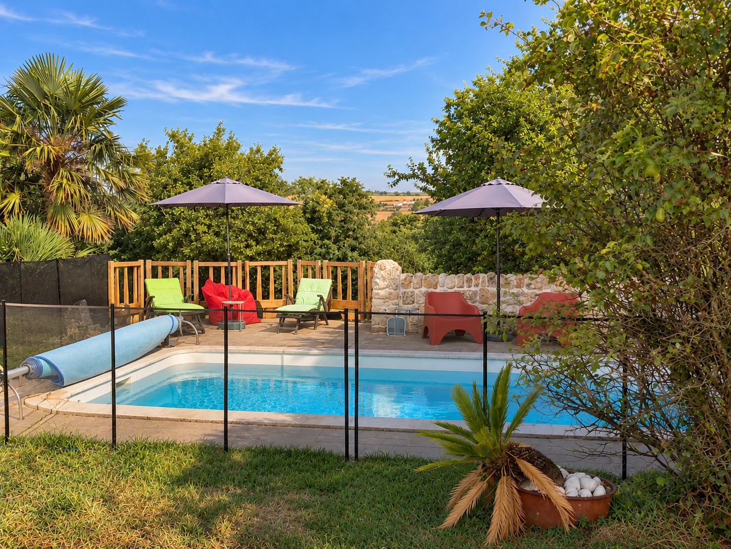 Une piscine bordée d’un muret en pierre et entourée d’une clôture en bois. Des chaises et des parasols aux couleurs vives sont disposés autour de la piscine.