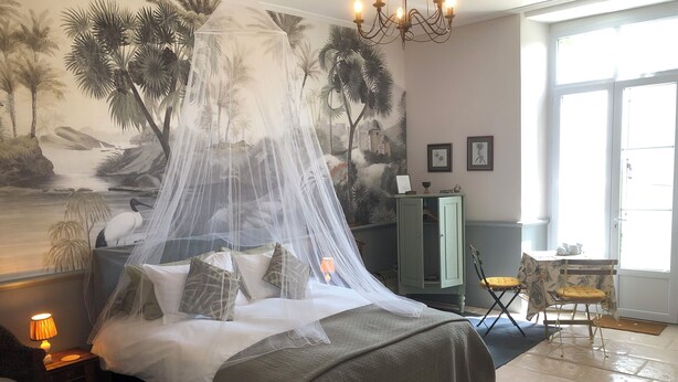 Ein Schlafzimmer mit einem Bett unter einem Baldachin, einem Nachttisch mit Lampe, einem grünen Schrank und einem Sessel mit gelber Kissenlehne.