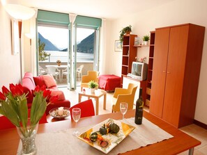 A cozy living room with a red sofa, a dining table with a plate of fruit, a bottle of wine, and a view of the sea through a balcony door.