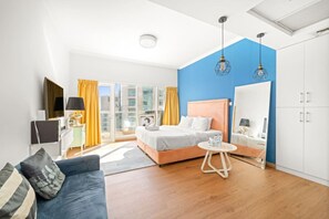 A modern hotel room with a bed, a sofa, a small table, a wardrobe, and a large window with curtains.