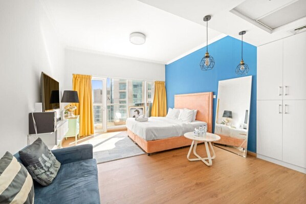 A modern hotel room with a bed, a sofa, a small table, a wardrobe, and a large window with curtains.