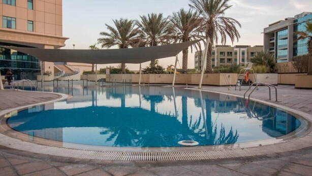A swimming pool with a shaded area, palm trees, and modern buildings in the background.