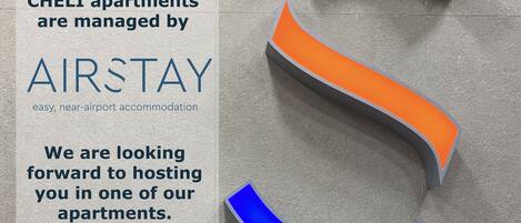 A sign for CHEL apartments managed by AIRSTAY, mentioning easy, near-airport accommodation and a look forward to hosting guests.