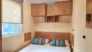 A compact bedroom with a bed, two pillows, a window, and built-in wooden cabinets. - Maia: Family bungalow (Canet de Mar)