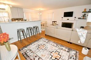 A modern living room with a sofa, a coffee table, and a rug. There is a kitchen area with a counter and stools. A flat-screen TV is mounted above a fireplace.