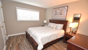 A bedroom with a wooden floor, a bed with a patterned bedspread, a nightstand with a lamp, and a wall-mounted artwork. - Condo at the University of Notre Dame D3 (South Bend)